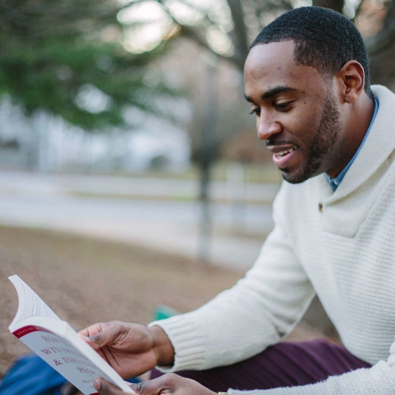 A smiling man in a cream sweater reading a book outdoors